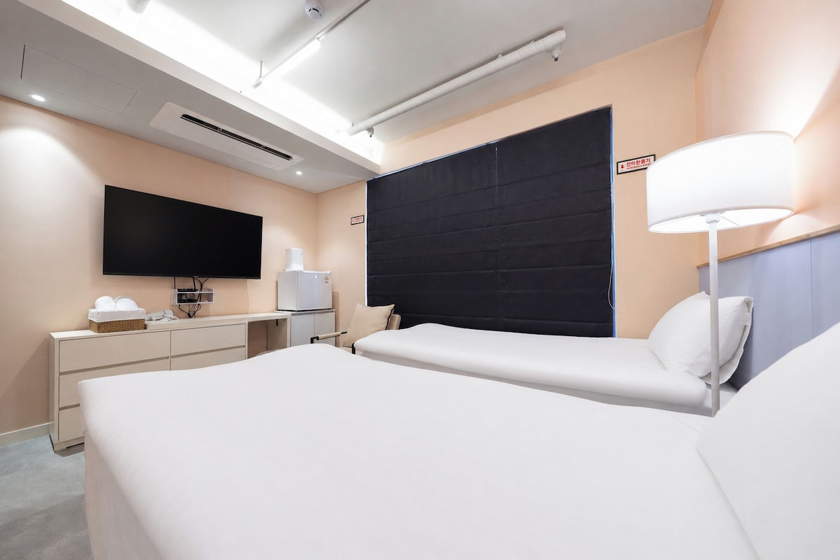 A hotel room with a bed, bedside table, lamp, flat-screen TV, and a large window with blackout curtains.