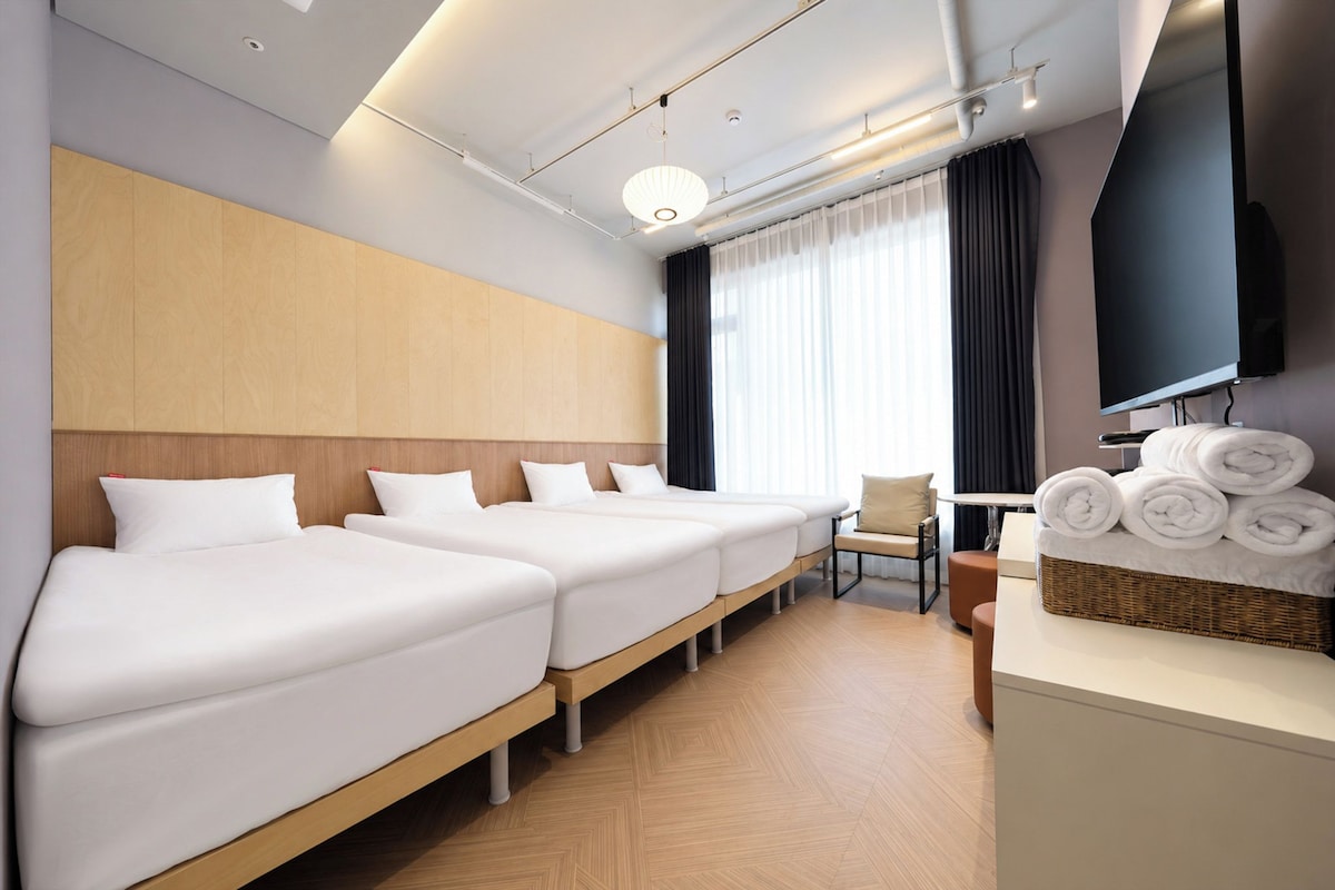 A hotel room with three beds, a flat-screen TV, a chair, and a basket of towels.