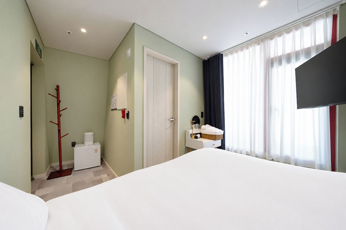 A hotel room with a bed, a desk with a chair, a TV, a coat rack, and a radiator.