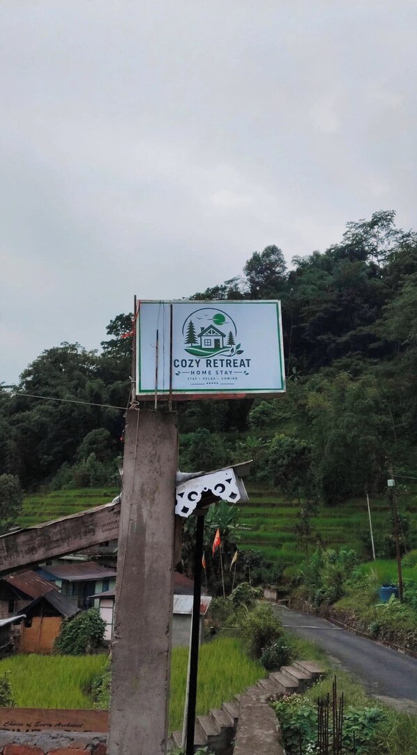A sign for a cozy retreat with a house and trees logo, surrounded by lush greenery and a rural setting. - Cozy Retreat Homestay mountain view homestay in Bijonbari-Darjeeling| Organic. (Goke)