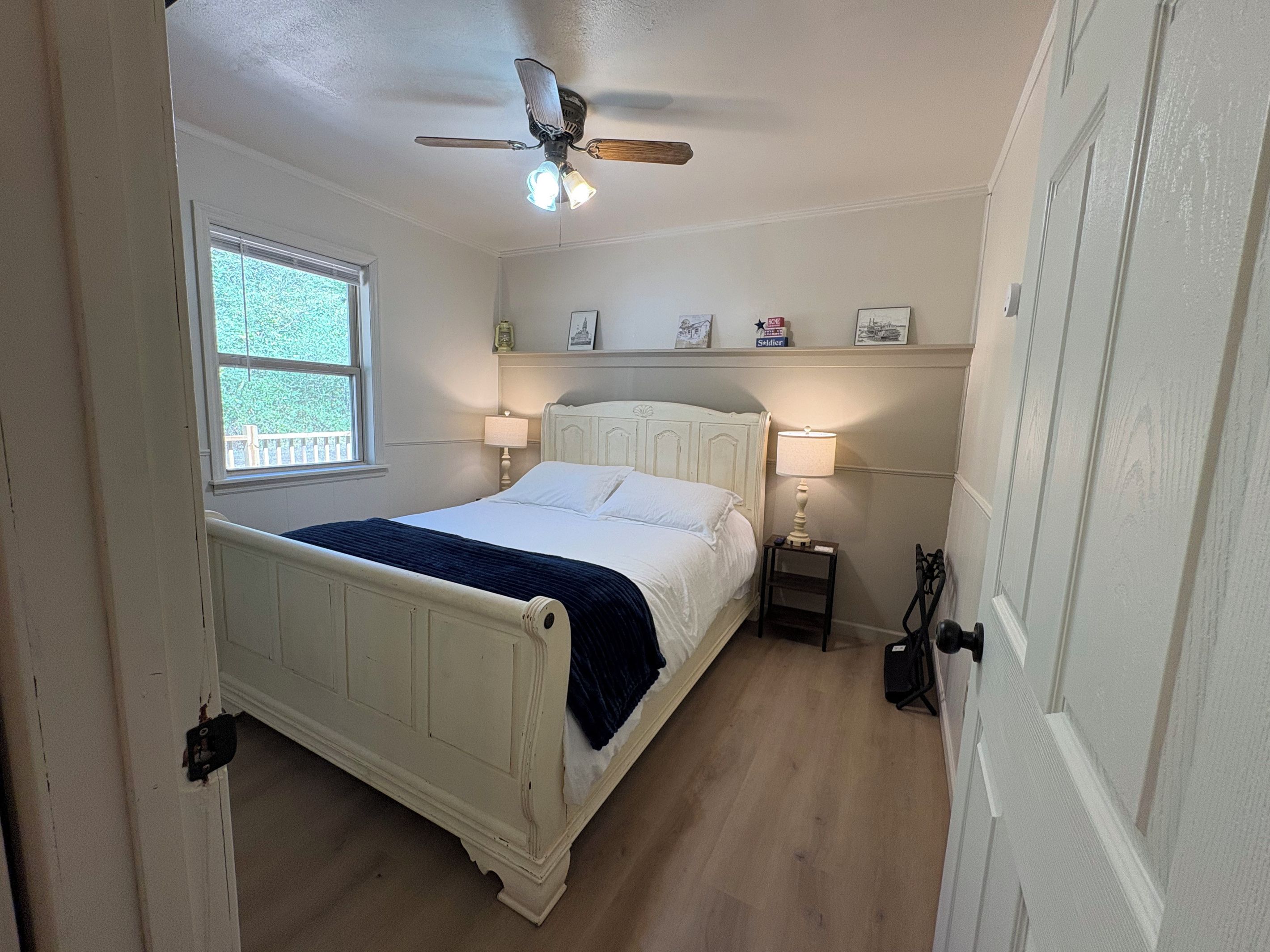 Un dormitorio con una cama individual, ventilador de techo, ventana y mesita de noche con lámpara.