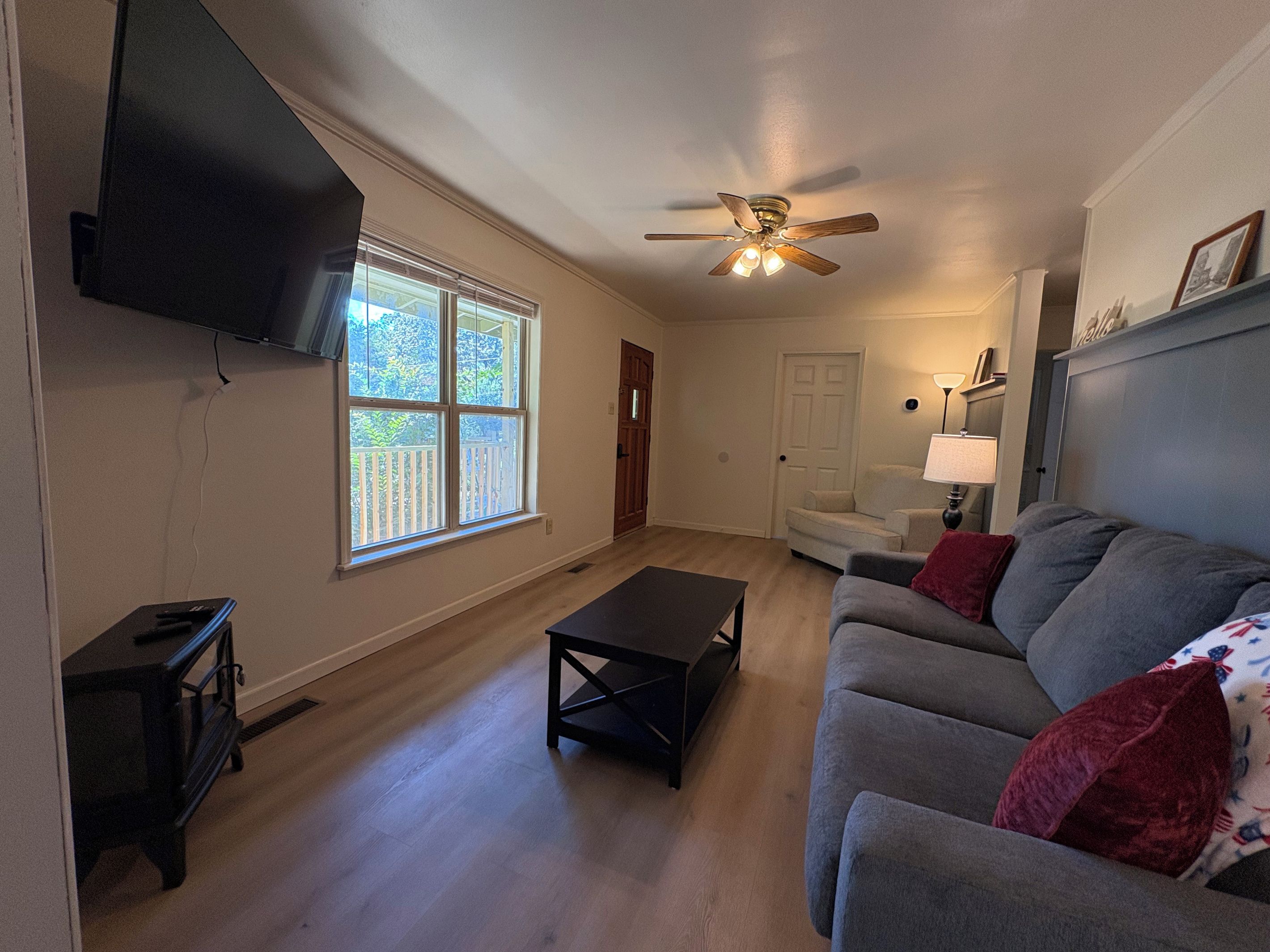 A living room with a flat-screen TV mounted on the wall, a ceiling fan, a window with a view of a deck, a grey sofa, and a small table.