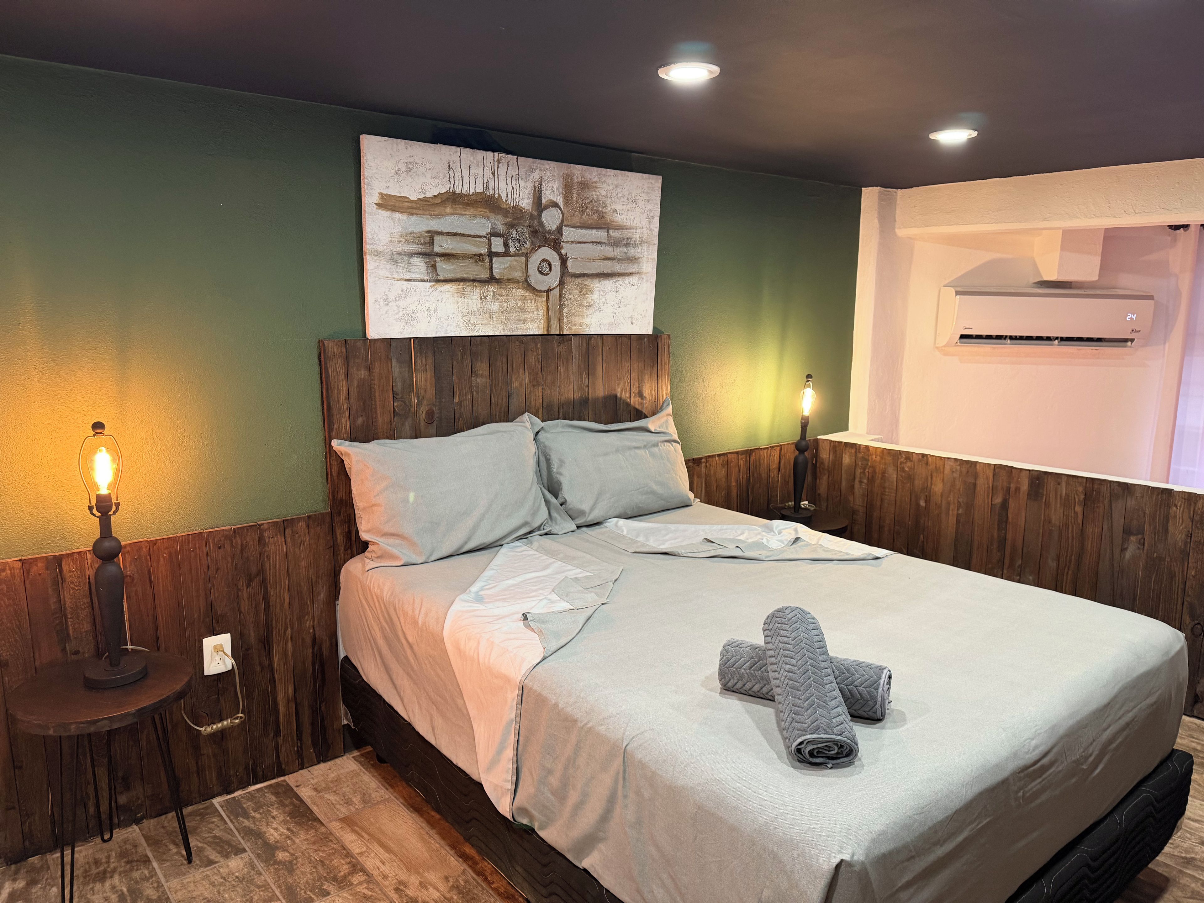 Un dormitorio con cabecera de madera, cama con ropa de cama gris, aire acondicionado de pared y mesita de noche con lámpara.