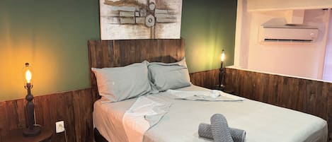 Un dormitorio con cabecera de madera, cama con ropa de cama gris, aire acondicionado de pared y mesita de noche con lámpara.
