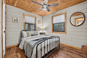 A bedroom with a bed, a ceiling fan, two windows with blinds, a bedside table with a lamp, and a mirror on the wall. - Woodland Hideaway | Hot Tub, Fire Pit & Fenced Yrd (Blue Ridge)
