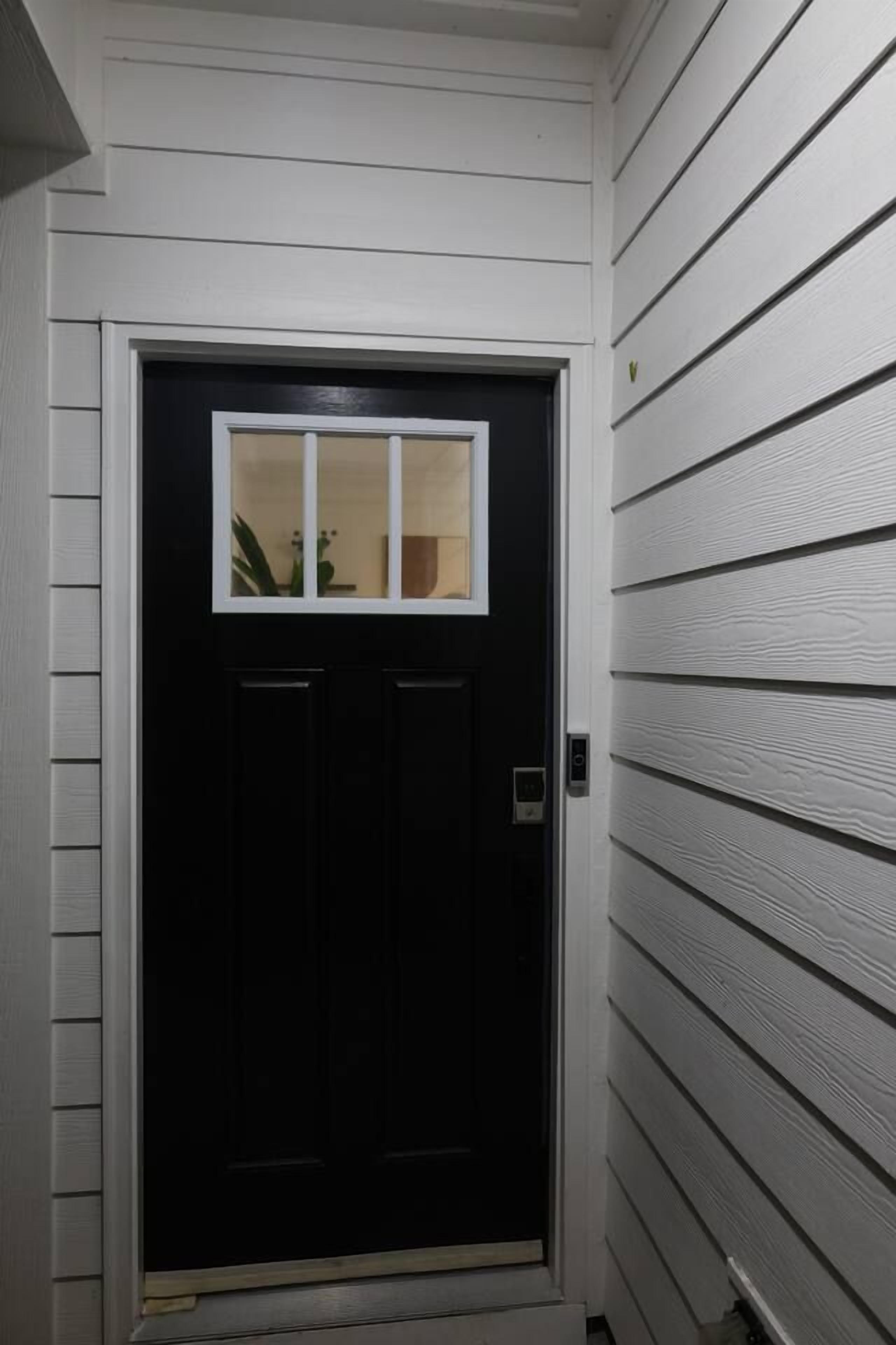 A black door with a small window, white siding on the walls, and a doorbell.