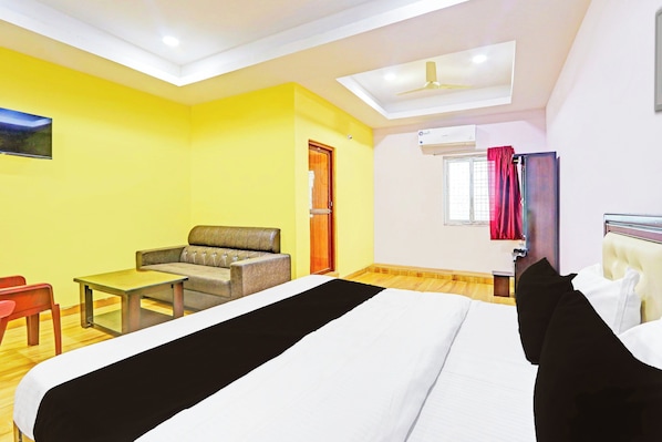 Habitación de hotel con cama, sofá, mesa, silla, ventilador de techo, unidad de aire acondicionado y ventana con cortinas.