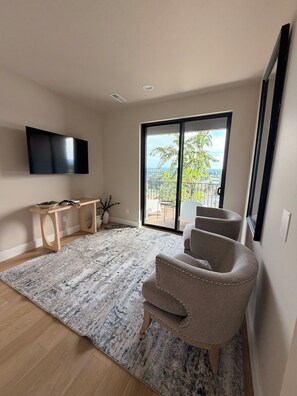 A modern living room with a flat-screen TV, a wooden coffee table, a patterned rug, a beige armchair, and a large sliding glass door leading to an outdoor balcony. - Perfect 3-bedroom townhome in gorgeous Vancouver with City Views and EV charger (Vancouver)