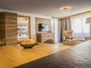 A modern hotel room with a wooden dining table, a television mounted on the wall, a comfortable armchair, and a large window with curtains. - Arcade Appartement/Fewo 4-Bettwohnung Nr. 115C02 by Interhome (Saas-Fee)