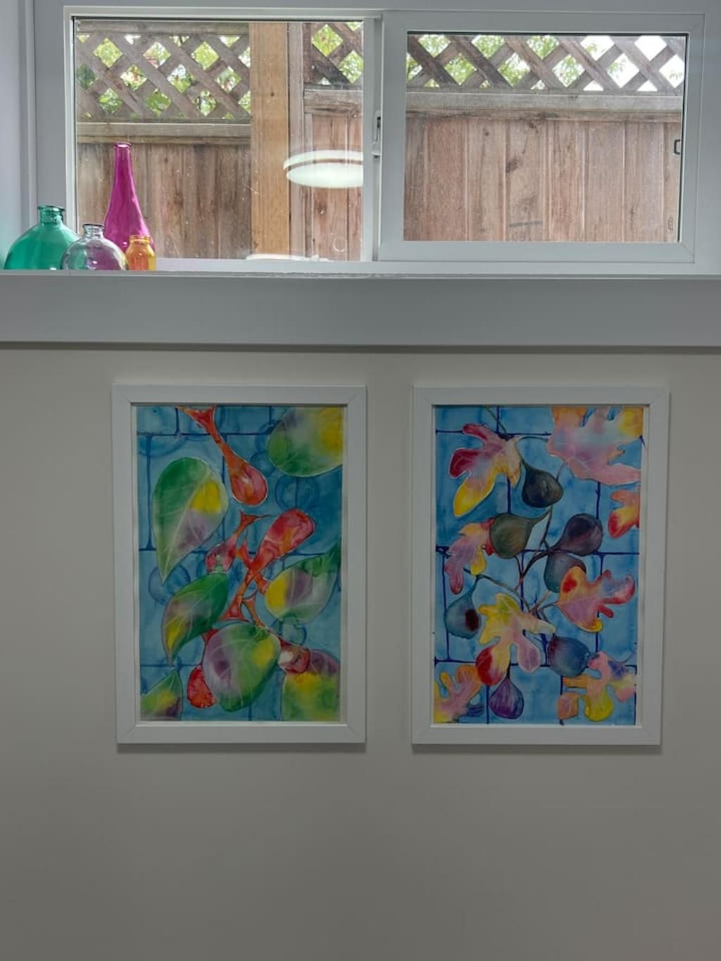Two framed paintings on a wall, a window with a view of a wooden fence and a hanging light fixture.