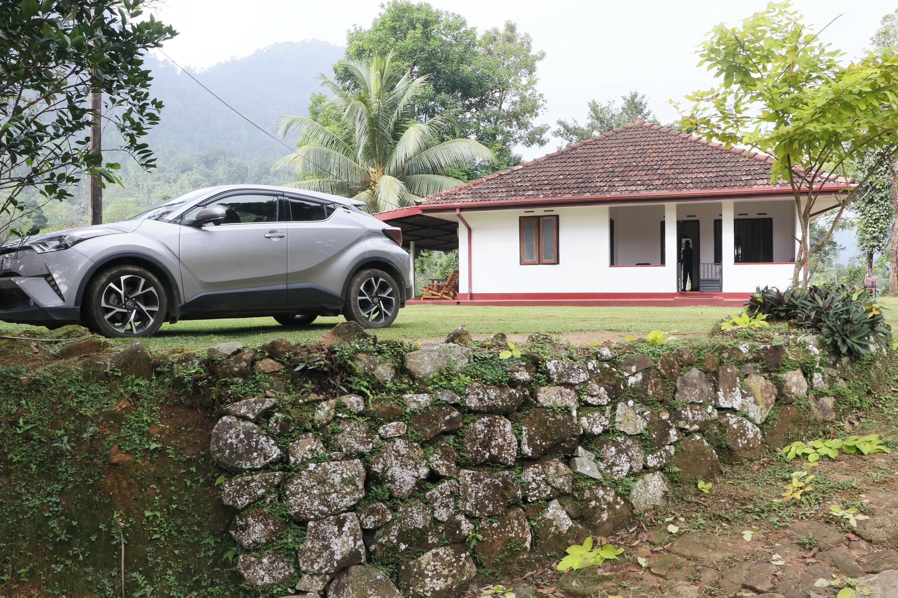 Um muro de pedra, uma casa com telhado vermelho e um carro estacionado.