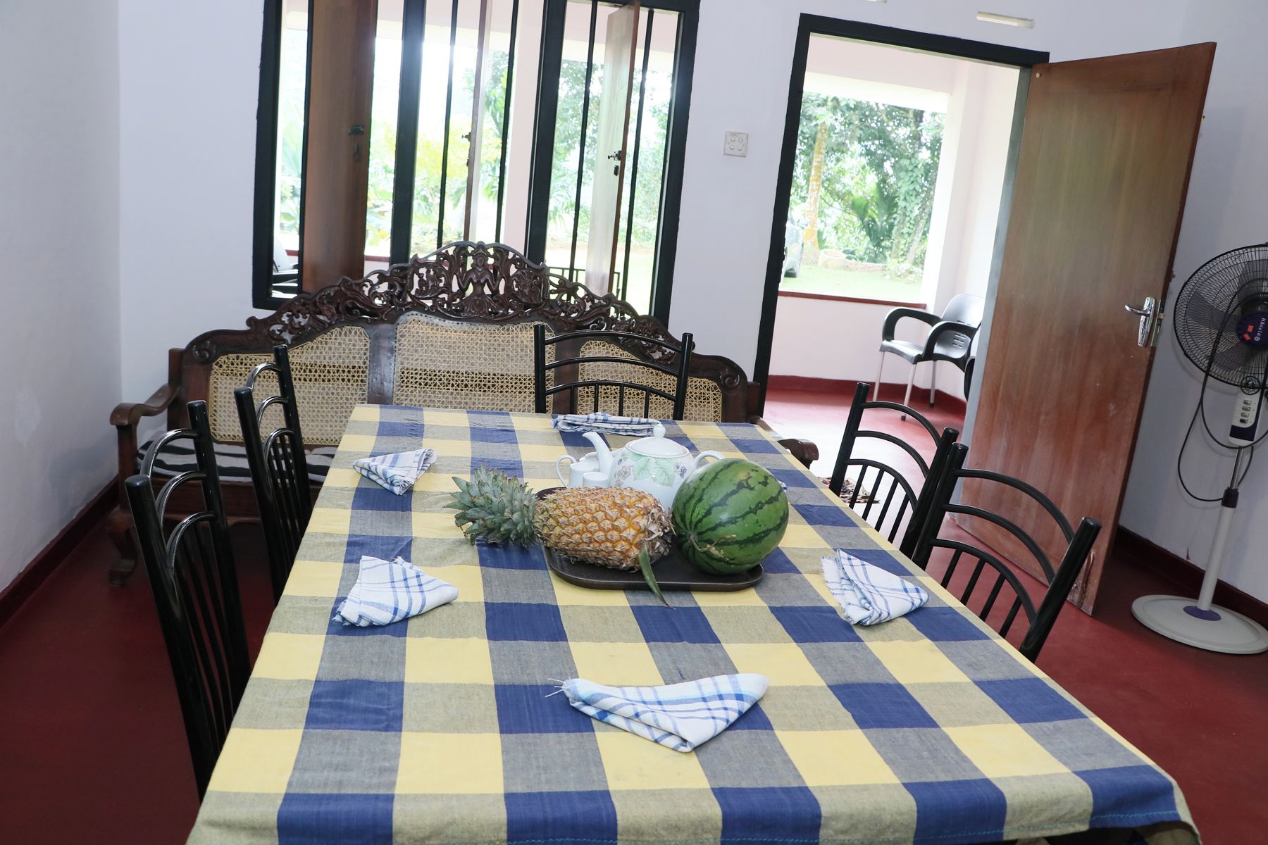 Uma área de refeições com toalha xadrez, abacaxi, melancia e ventilador.