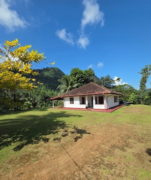 A single-story house with a red roof and white walls, surrounded by a grassy area and trees.