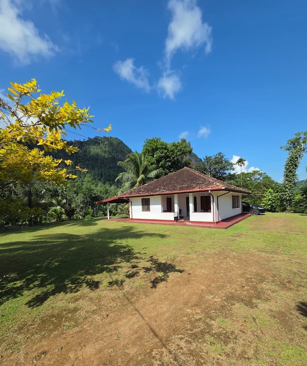 A single-story house with a red roof and white walls, surrounded by a grassy area and trees.