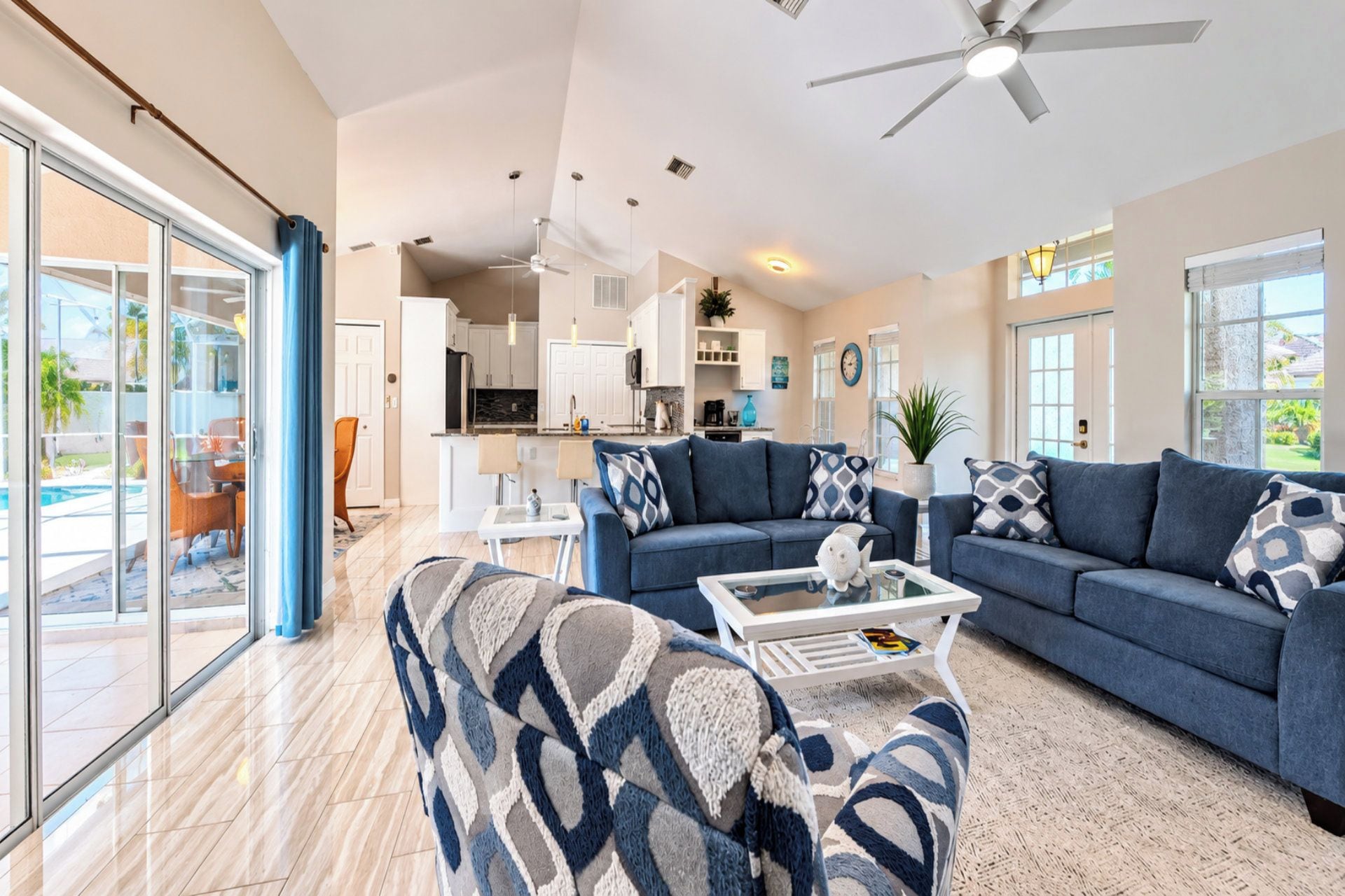 A spacious living room with a large blue sofa set, a coffee table, and a view of the kitchen and outdoor area.