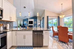 A modern kitchen with a central island, stainless steel appliances, and a dining area with a view of the outdoors. - VRCC Villa The Owl’s Nest (Cape Coral)