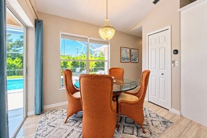 A dining area with a glass table, wicker chairs, a chandelier, and a view of a pool through a window. - VRCC Villa The Owl’s Nest (Cape Coral)