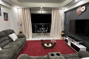 A modern living room with a grey sofa, a red rug, a flat-screen TV, and a balcony with a table and chairs. - 2-beds apartment in Westlands near GTC Free airport pick up with fitness access (Nairobi)