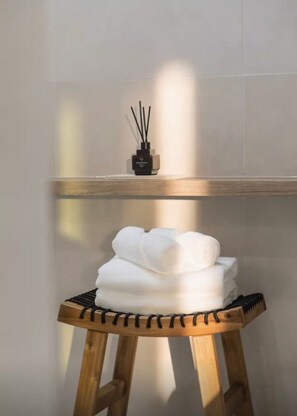 Ein Holzschemel mit einem weißen Handtuch darauf, eine schwarze Flasche mit einem Reed-Diffusor und ein Holzregal an einer gefliesten Wand.