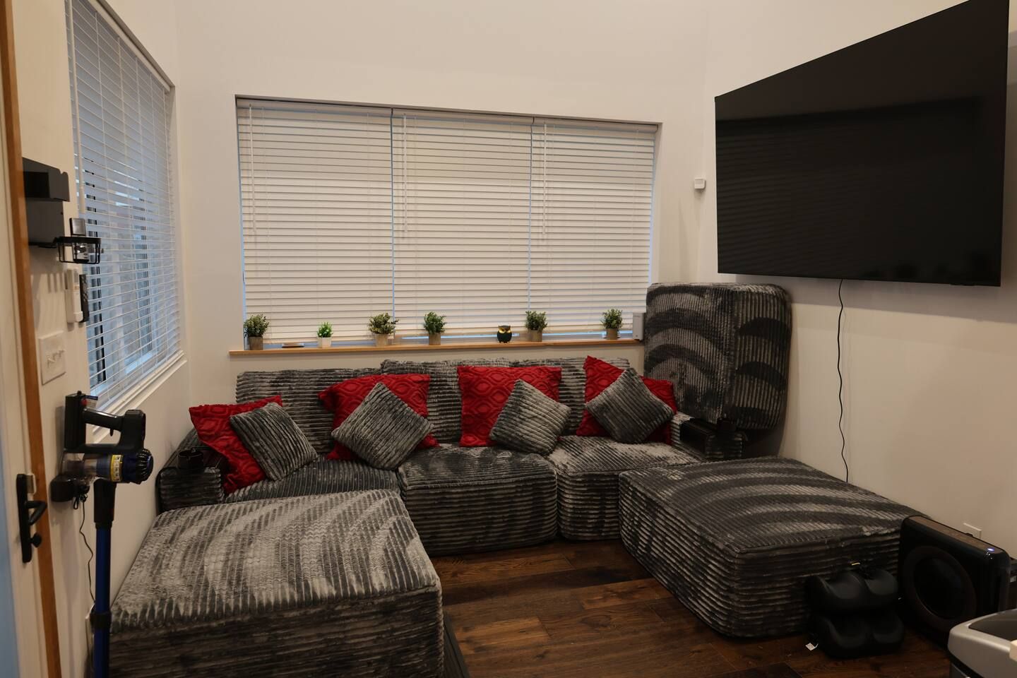 Ein Wohnzimmer mit einer Eckcouch, einem Flachbildfernseher und einem Fenster mit Jalousien.