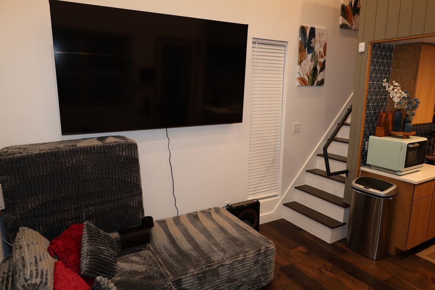 A living room with a flat-screen TV mounted on the wall, a gray sofa with red cushions, a staircase leading to a kitchen area with a microwave and a trash bin.