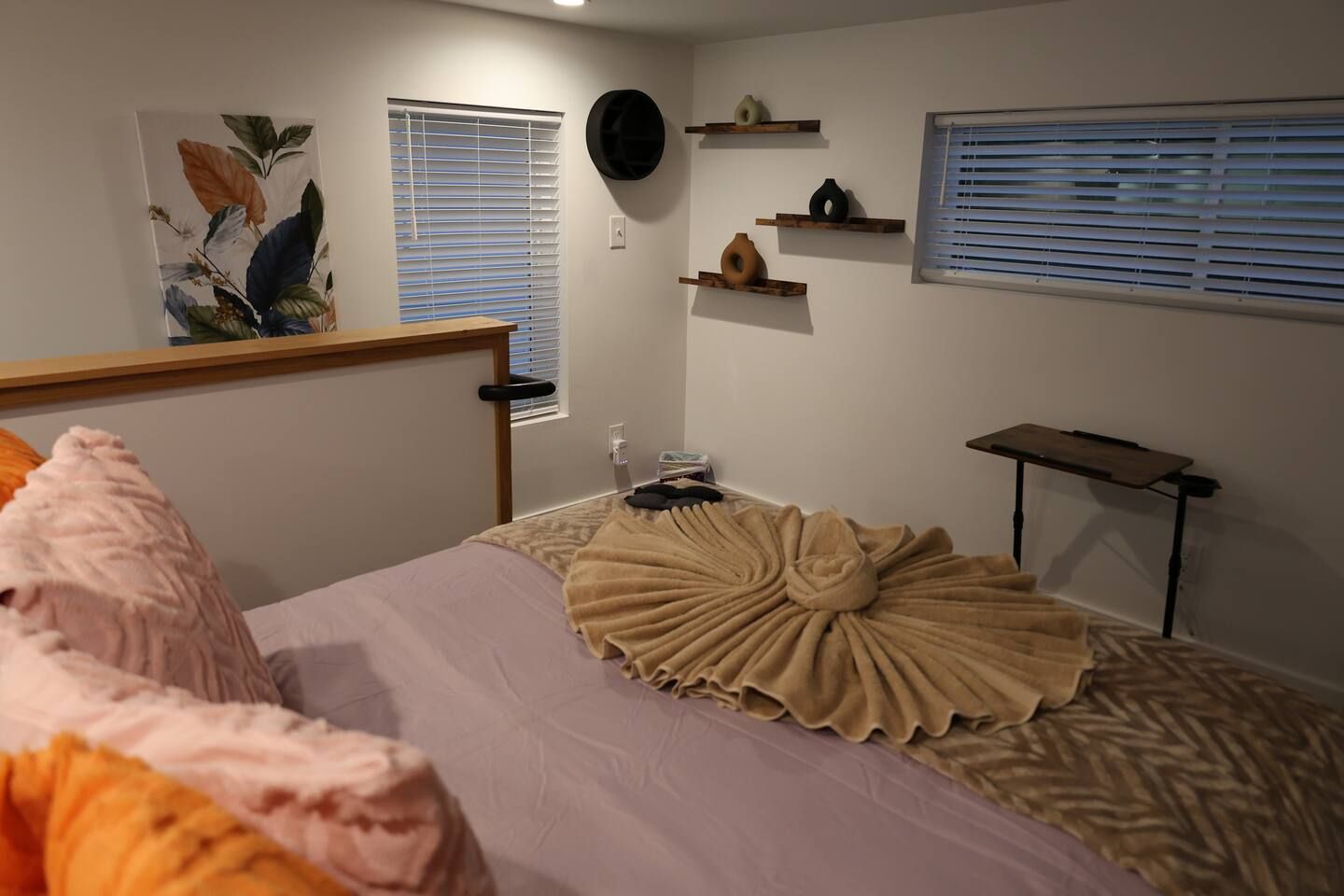 A bedroom with a bed, a bedside table, a wall-mounted shelf with decorative items, and a window with blinds.