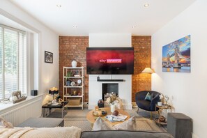 A living room with a fireplace, a television mounted above it, a bookshelf, and a comfortable seating area. - Home cinema and super king bed - Hampstead (Greater London)