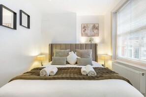 A hotel room with a large bed, two bedside lamps, a window with blinds, and a framed picture on the wall. - Home cinema and super king bed - Hampstead (Greater London)