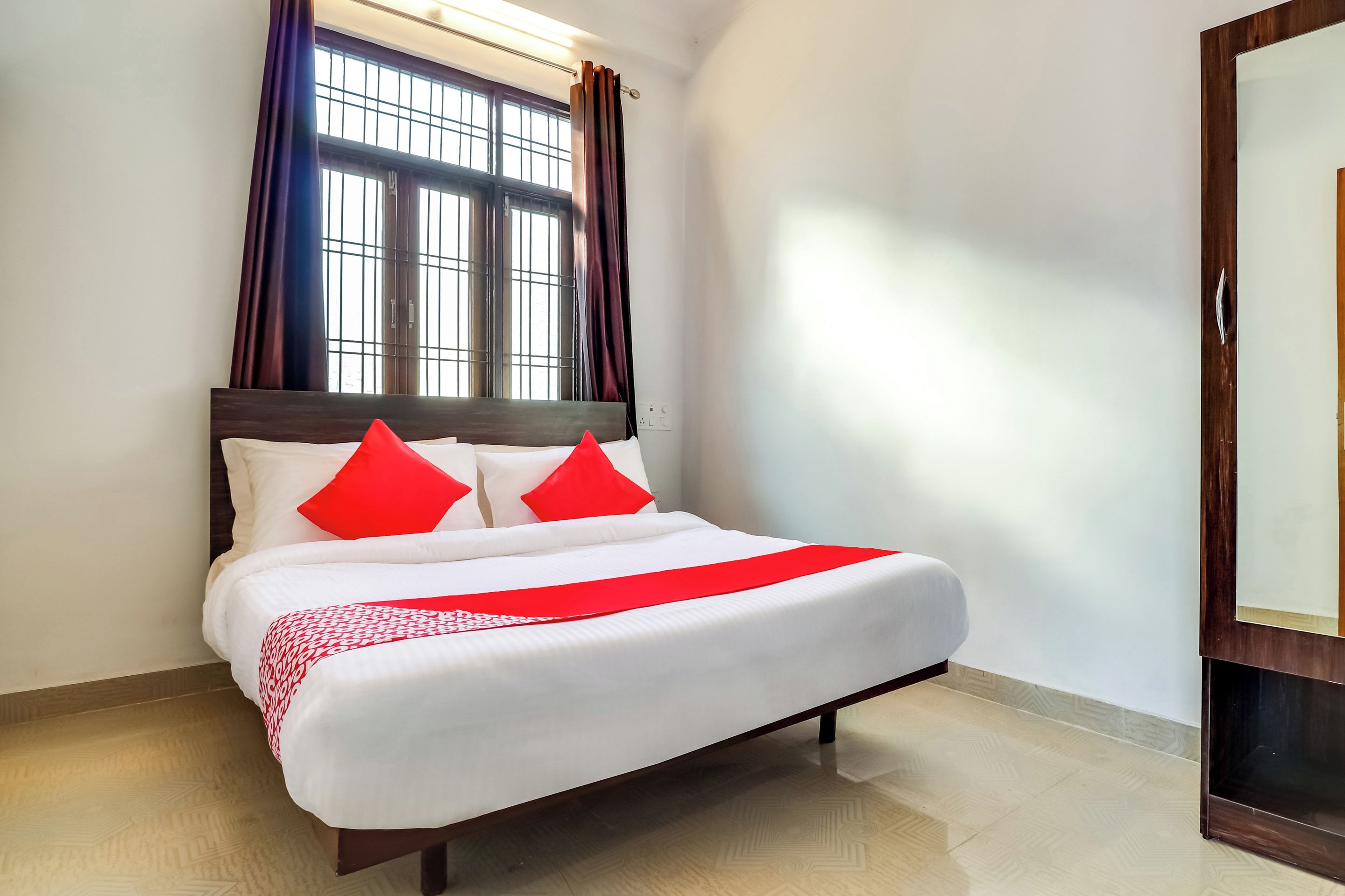 A neatly made bed with white linens and red accents, a window with curtains, and a wooden floor.