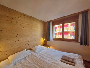 Habitación de hotel con cabecera de madera, cama con ropa blanca, ventana con cortinas y vista a un edificio con balcones.