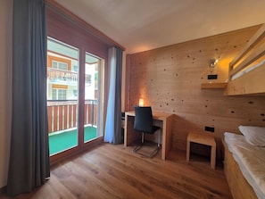 A room with a wooden floor, a desk with a chair, a bed, and a balcony with a view of a building. - Dominic A1 Appartement/Fewo Appartement/Fewo by Interhome (Saas-Fee)