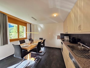 A modern kitchen with dark flooring, a dining area with a wooden table and chairs, and a view of greenery through the window. - Dominic A1 Appartement/Fewo Appartement/Fewo by Interhome (Saas-Fee)