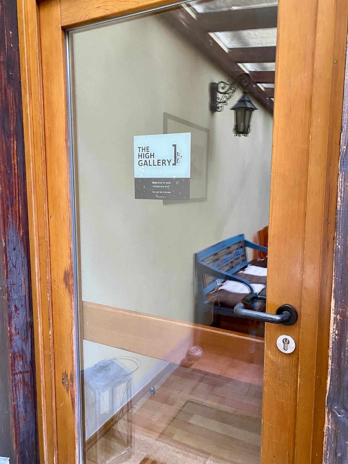 A wooden door with a glass panel, a sign reading 'The High Gallery,' and a glimpse of a room with a bed and a blue chair.
