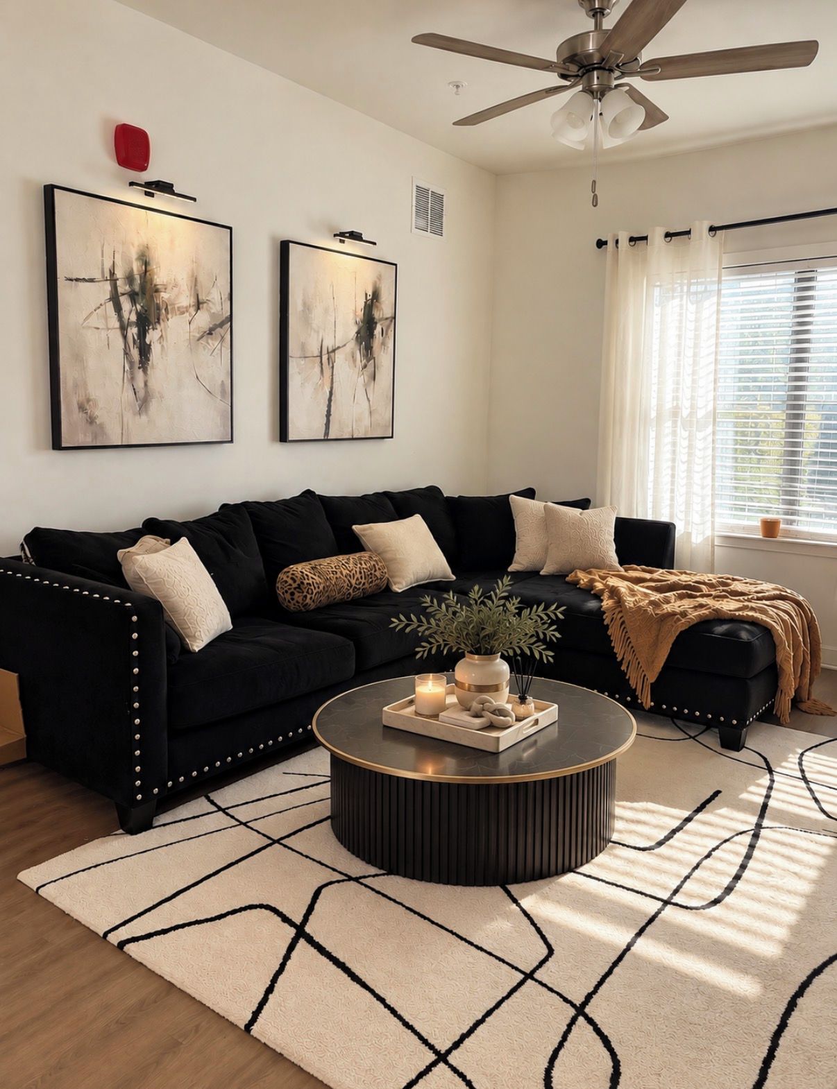 Un salon moderne avec un canapé noir, deux tableaux abstraits, un ventilateur de plafond et une table basse ronde agrémentée d’objets décoratifs.