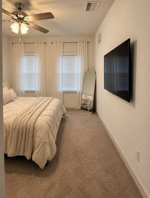 Une chambre à coucher comprenant un lit, un ventilateur de plafond, deux fenêtres équipées de stores et un téléviseur fixé au mur.