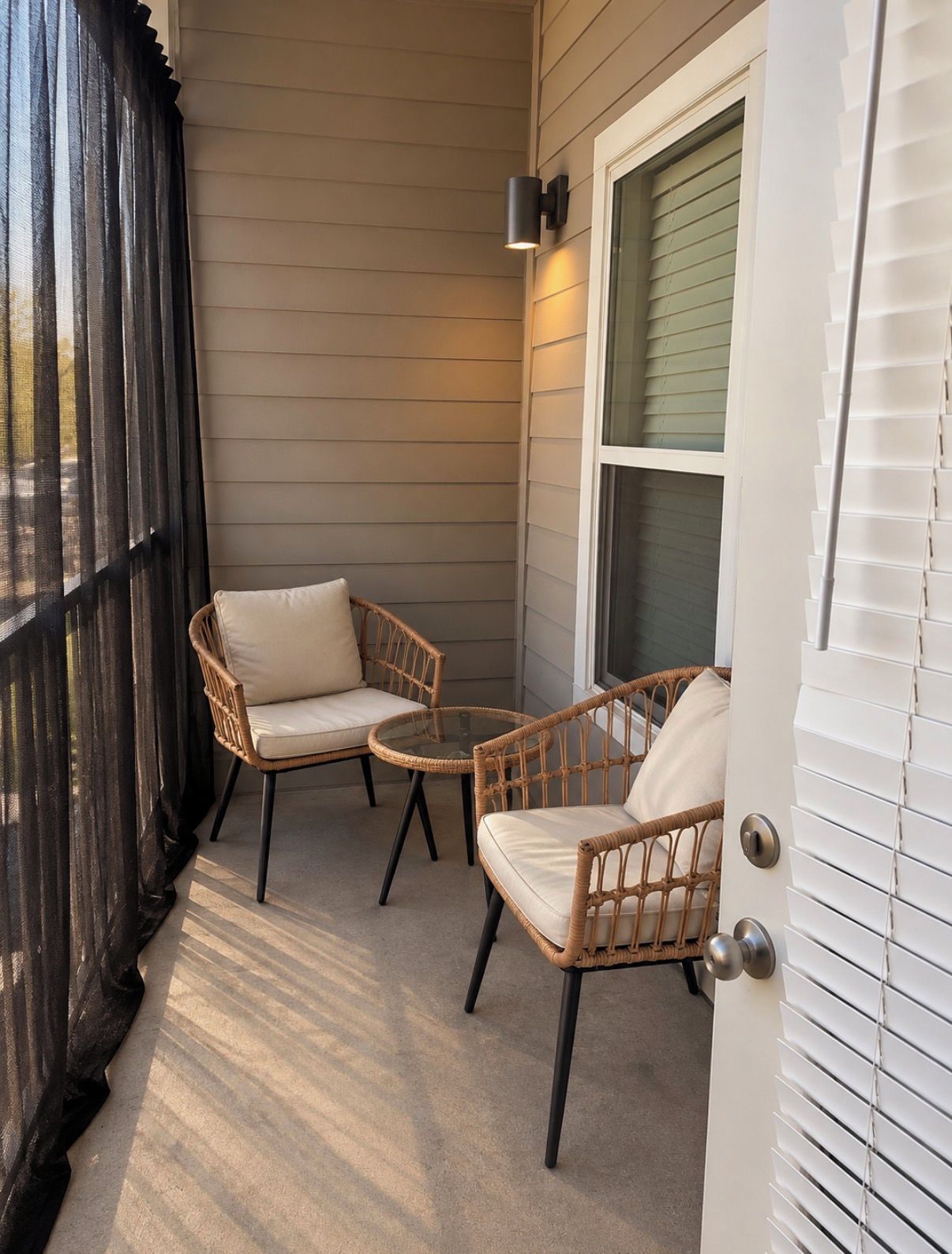 A covered porch with two wicker chairs and a small round table, a window with blinds, and a door with a handle.
