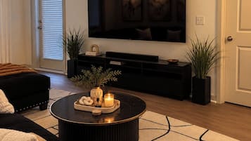 A modern living room with a black sofa, a round coffee table with decorative items, a large flat-screen TV mounted on the wall, and a patterned rug.