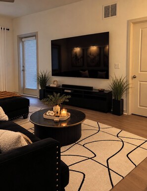 Un salon moderne avec un canapé noir, une table basse ronde agrémentée d’objets décoratifs, un grand téléviseur à écran plat fixé au mur et un tapis à motifs.