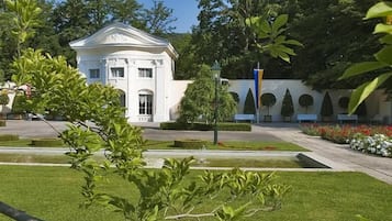 Ein weißes Gebäude im klassischen Stil, umgeben von einem gepflegten Garten mit geschnittenen Hecken und blauem Himmel.