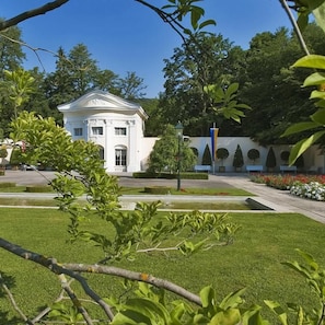 A white building with a classical architectural style, surrounded by a well-maintained garden with trimmed hedges and a clear sky.
