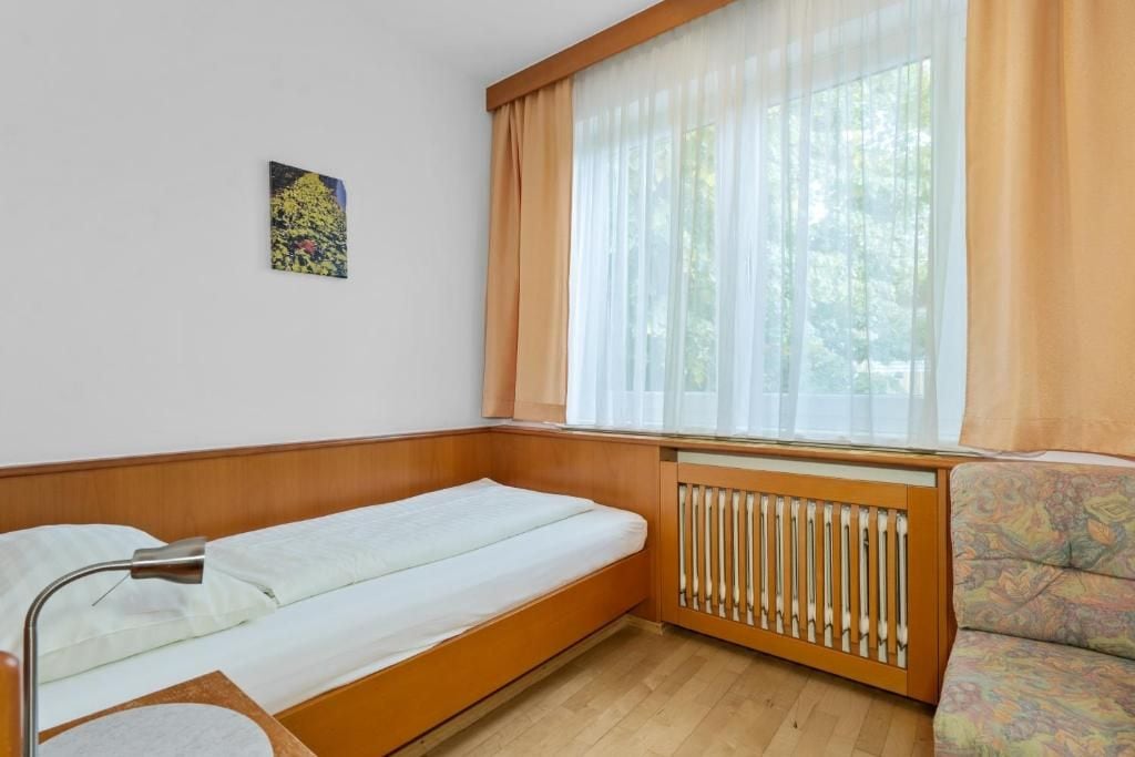 Una cama individual con cabecera, una lámpara de noche, un radiador y una ventana con cortinas traslúcidas.