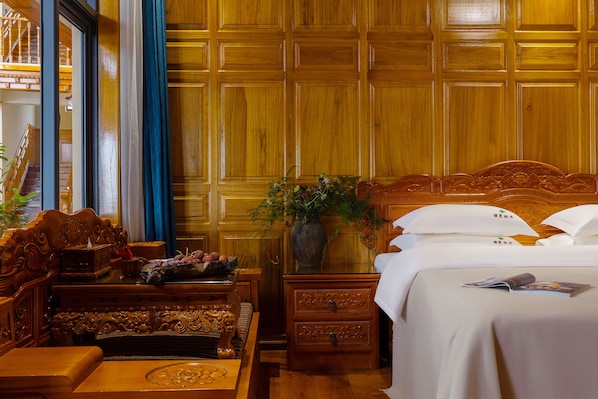 A hotel room with a wooden paneled wall, a bed with white linens, a wooden nightstand with a vase of flowers, a wooden bench, and a table with a decorative bowl.