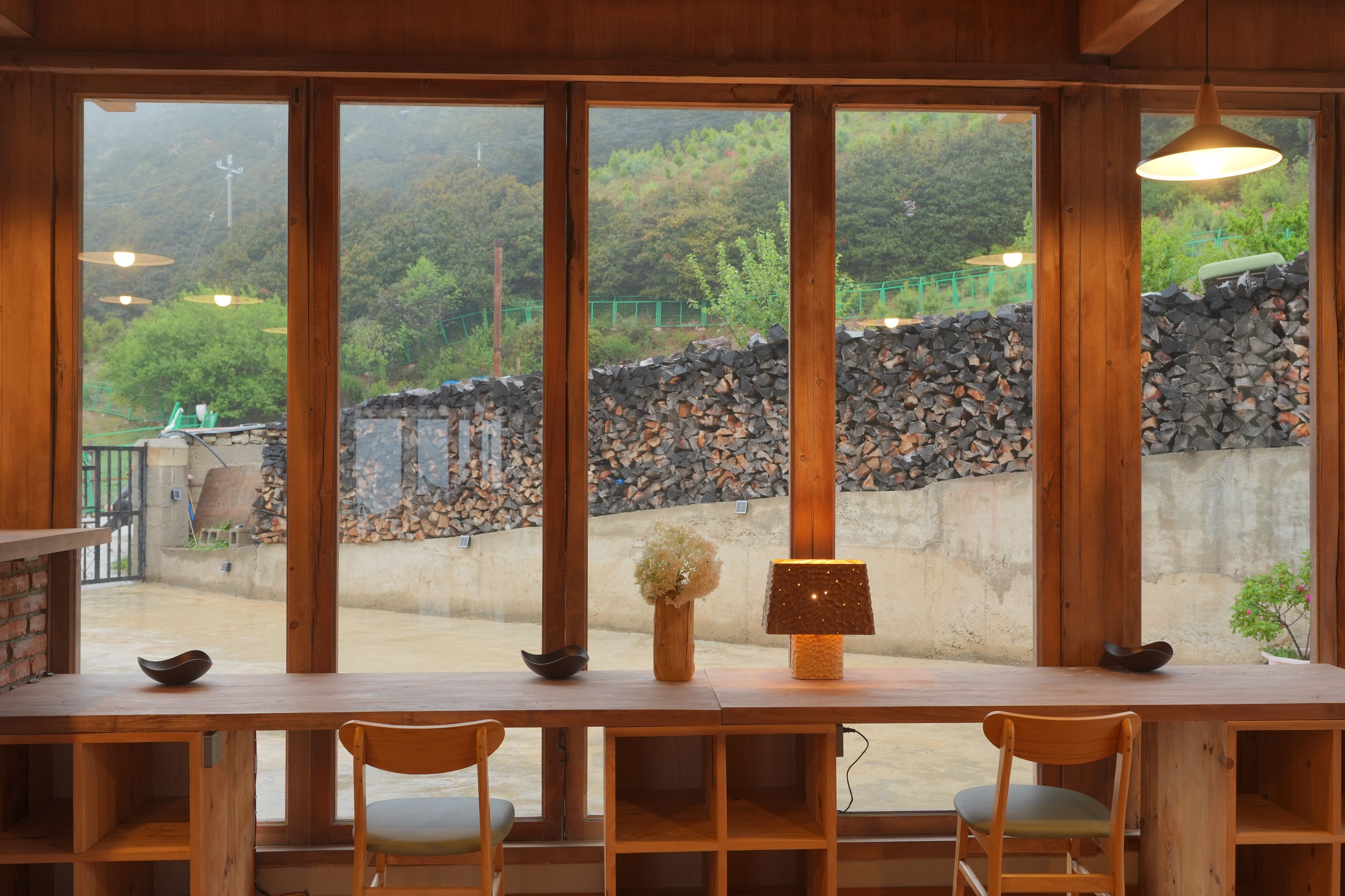 Interior de madera con ventanas amplias, una mesa con sillas y vistas a una pared de piedra y vegetación al exterior.