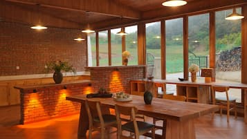 Een houten interieur met een bakstenen muur, een houten tafel en stoelen, en grote ramen met uitzicht op groen.