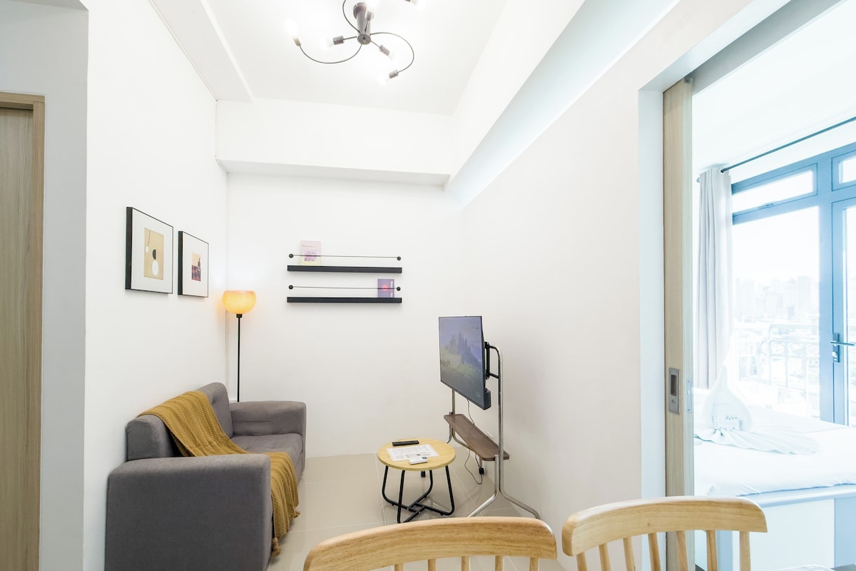 Una sala de estar moderna con un sofá gris, una pequeña mesa con sillas, un televisor de pantalla plana montado sobre un soporte y un ventilador de techo.