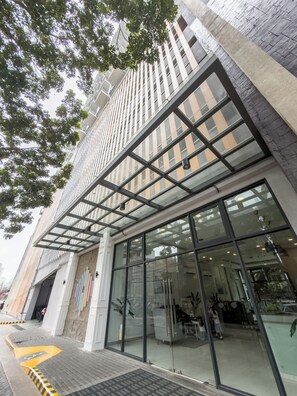 A modern building with large glass windows, a metal framework, and a paved entrance area.