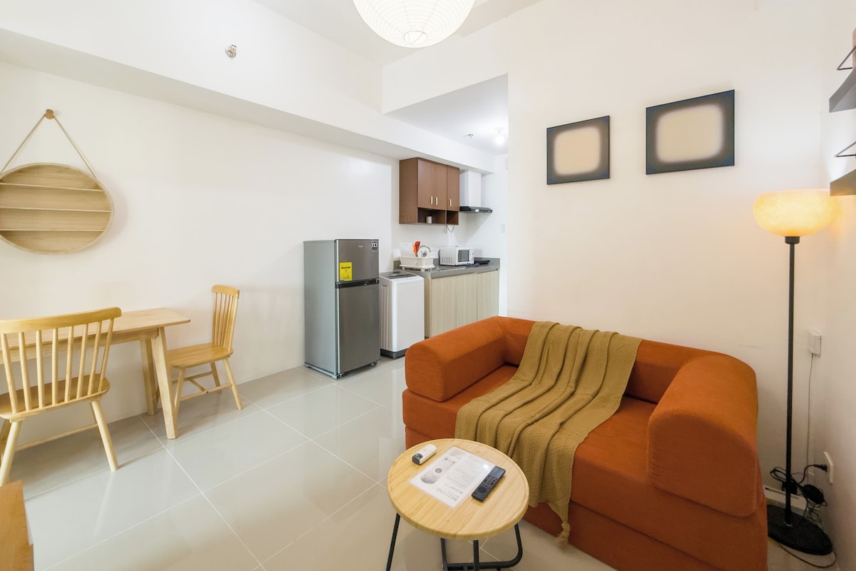 Una sala moderna con un sofá, una mesa de comedor con sillas, una mesita redonda con un control remoto y una zona de cocina al fondo.