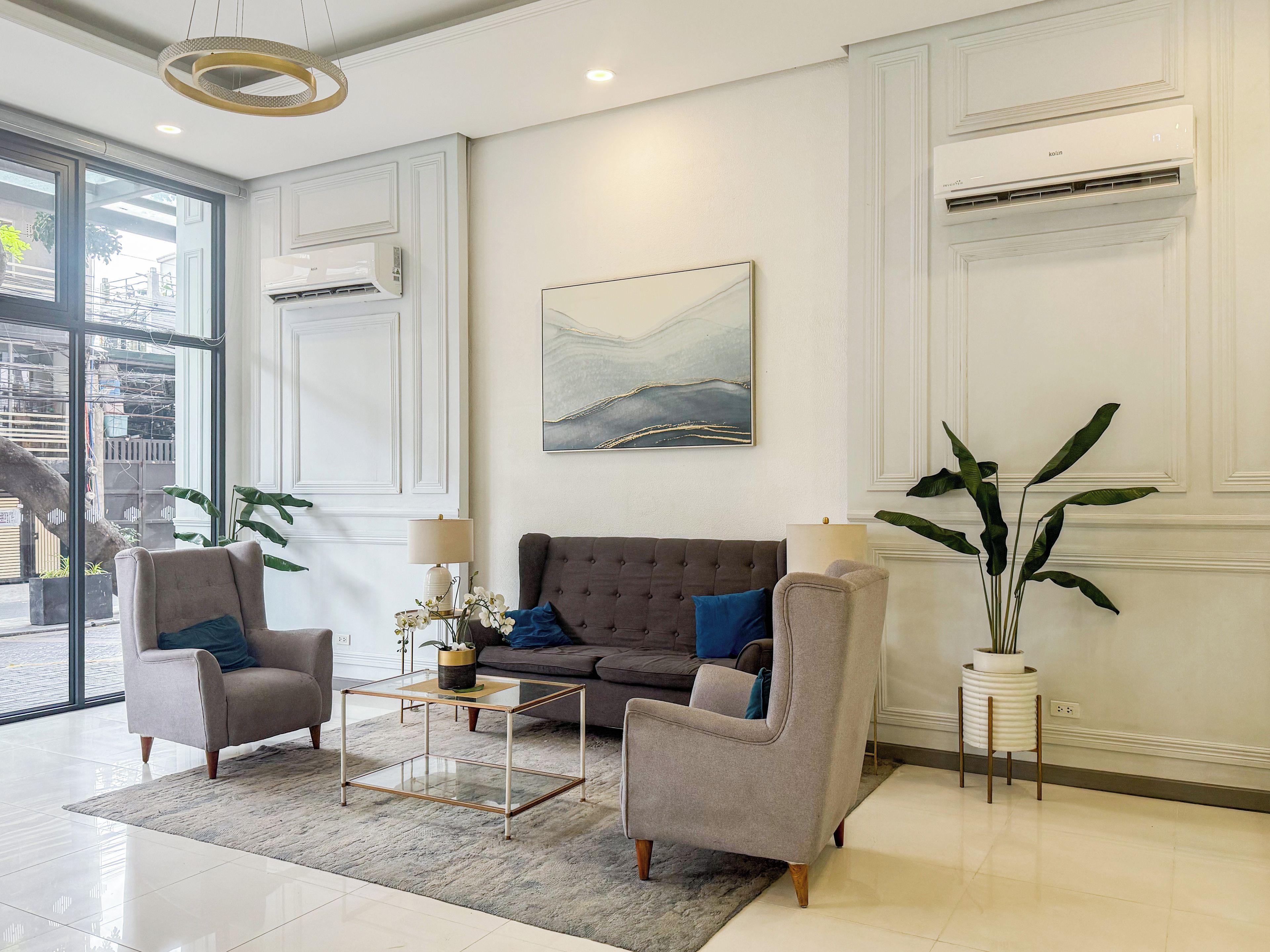A modern living room with a grey sofa, two armchairs, a glass coffee table, and a large window offering a view of the street.