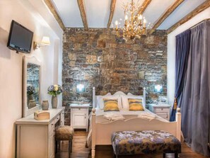 A bedroom with a stone wall, a bed with patterned pillows, a nightstand, a desk with a mirror, a television, and a chandelier. - Peaceful Pool Villa in Crete Countryside (Apokoronas)
