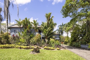 A residential property with a well-maintained lawn, various tropical plants, and a two-story house with a balcony. - Peaceful 2-bedroom in Innisfail Cassowary Coast (Belvedere)
